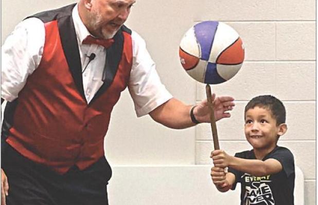 Juggler Richard Holmgren gets some assistance from Dexter Wiebe who balances a spinning basketball on a stick during the “Flying Debris” program held at the Scott County Library on Tuesday morning. (Record Photo)