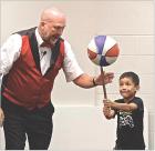 Juggler Richard Holmgren gets some assistance from Dexter Wiebe who balances a spinning basketball on a stick during the “Flying Debris” program held at the Scott County Library on Tuesday morning. (Record Photo)